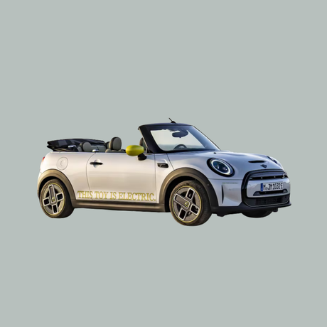 Een witte Mini Cooper cabriolet met gele spiegels en de zin THIS TOY IS ELECTRIC op de zijkant, geparkeerd op een effen grijze achtergrond.
