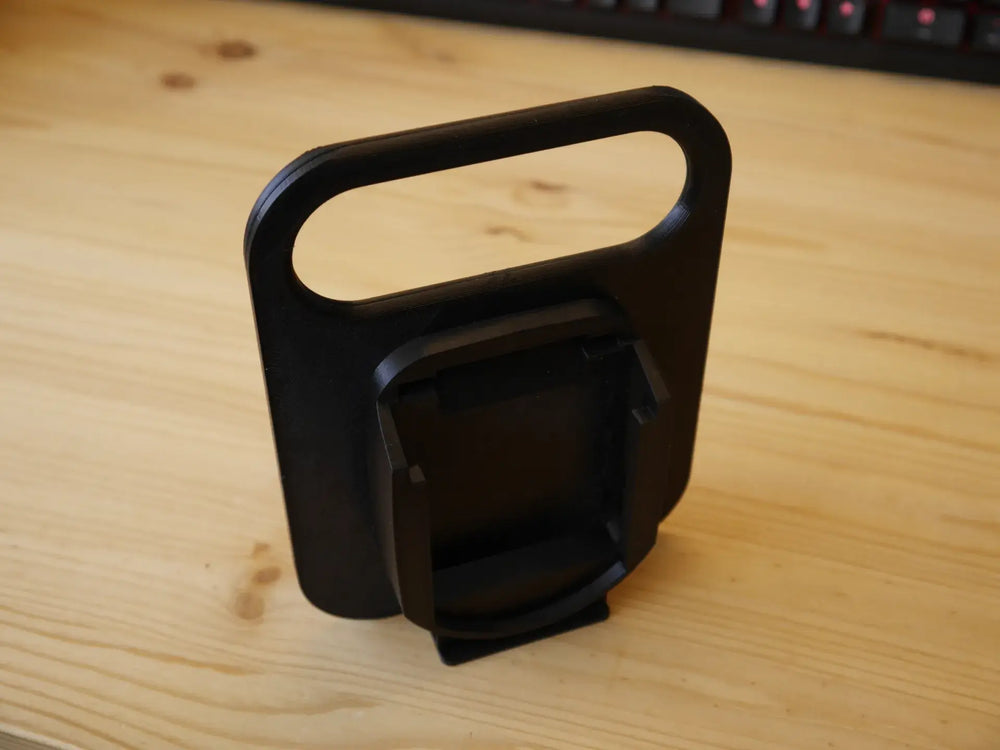 Een Makita LXT 18V BL1830-BL1890 Accuhouder & Transport Drager | 3D Geprint, zwart plastic met een rechthoekig handvat en schuine houder, staat op een licht houten bureau met een toetsenbord op de achtergrond.