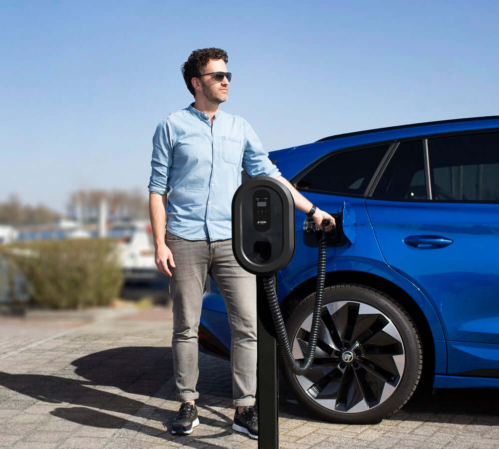 Een man met een zonnebril en een lichtblauw shirt laadt een blauwe elektrische auto op bij een Ratio EV laadstation met Montagepaal Dubbel voor Ratio EV Laadstation in de parkeerplaatsen op een zonnige dag.