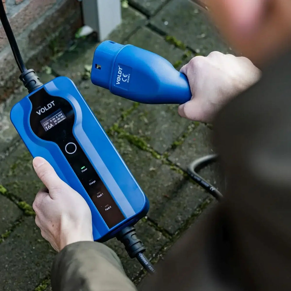 Een persoon houdt een Voldt Type 2 CEE 16A Mobiele Laadkabel (1-Fase, 3,7kW) vast, klaar om zijn elektrische auto buiten op te laden op een verharde ondergrond in de buurt van een bakstenen muur.
