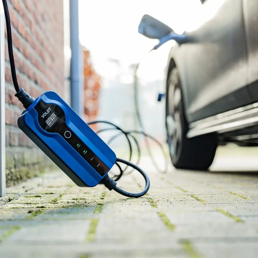 Een close-up van een zwarte elektrische auto-oplader met de Voldt Type 2 CEE 16A Mobiele Laadkabel 1-fase instelbaar 3,7kW (Nederlands product, 3 jaar garantie) bij een bakstenen muur, met het apparaat en de snoeren zichtbaar op de grond.