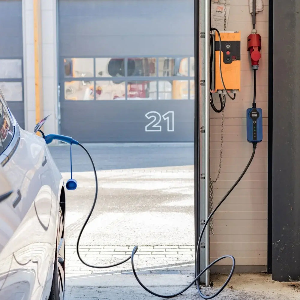 Een witte elektrische auto staat geparkeerd in een garage en wordt opgeladen met een 🇳🇱 Voldt 11kW 3-fase mobiele EV-lader (NL product, 3 jaar garantie). Zonlicht stroomt door de open deur en onthult een ander gebouw met nummer 21.