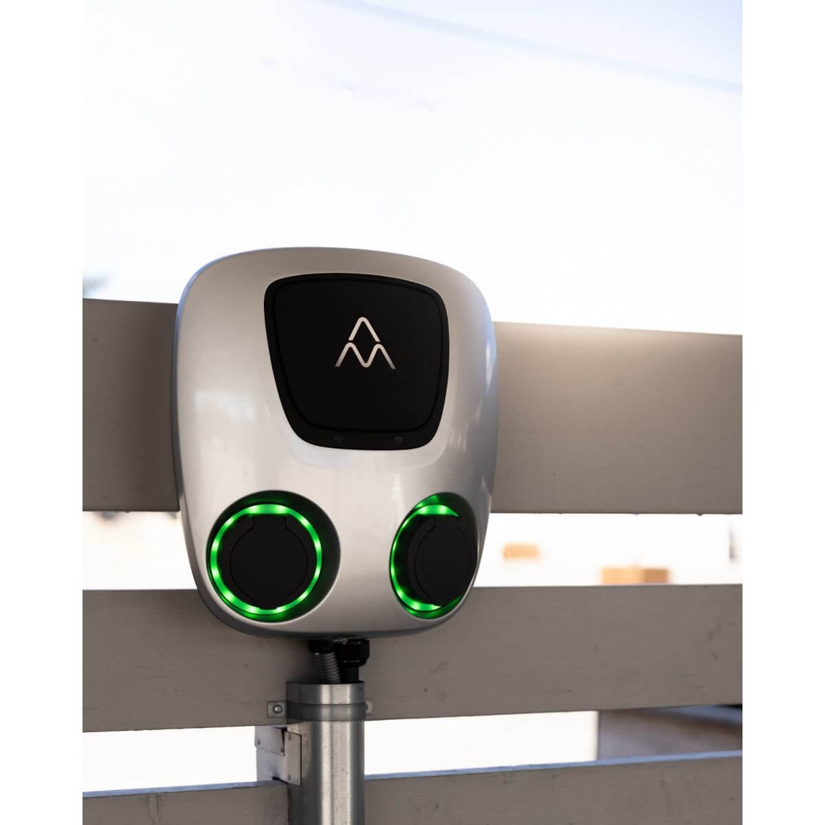 De Laadstation Charge Amps Aura met Sim 2x22 kW 3-fase 32A bi-directioneel voertuig naar net heeft twee ronde groen verlichte poorten, aan de muur gemonteerd op een wit houten hek met een centraal zwart display en wit logo.