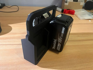 Een Makita Accuhouder & Transport Drager | 3D Geprint | Zwart is gemonteerd op hout, houdt een Makita 18V 5.0Ah accu stevig vast, met assorti gereedschap en dozen op de achtergrond.