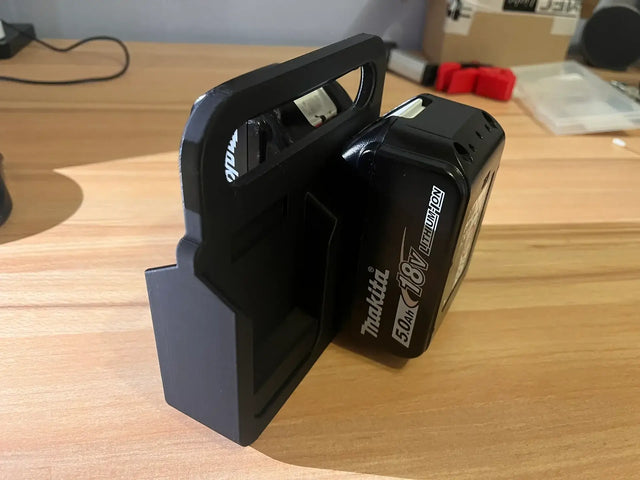 Een Makita Accuhouder & Transport Drager | 3D Geprint | Zwart is gemonteerd op hout, houdt een Makita 18V 5.0Ah accu stevig vast, met assorti gereedschap en dozen op de achtergrond.