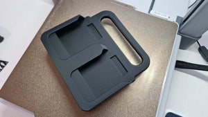 Een Makita Accuhouder & Transport Drager | 3D Geprint | Zwart rust op een gesloten, metallic gouden laptop op een wit bureau, met kabels en kantoorapparatuur zichtbaar op de achtergrond.