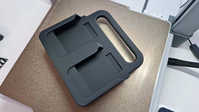 Een Makita Accuhouder & Transport Drager | 3D Geprint | Zwart rust op een gesloten, metallic gouden laptop op een wit bureau, met kabels en kantoorapparatuur zichtbaar op de achtergrond.