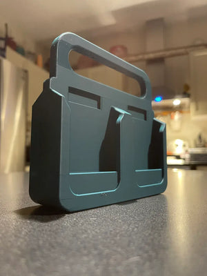 Een zwarte 3D-geprinte Makita Accuhouder & Transportkar met een handvat en twee sleuven staat op een aanrecht, met vage apparaten op de achtergrond.