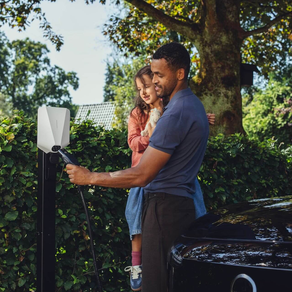 Een man houdt een jong meisje vast terwijl hij een oplaadkabel in een zwarte elektrische auto steekt op een buitenstation met groene bomen, genietend van het gemak van het Easee UP 22kW oplaadpunt elektrisch voertuig.