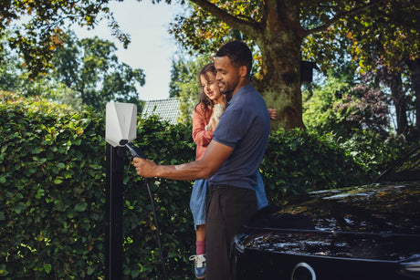 Een man houdt een jong meisje vast terwijl hij een oplaadkabel in een zwarte elektrische auto steekt op een buitenstation met groene bomen, genietend van het gemak van het Easee UP 22kW oplaadpunt elektrisch voertuig.